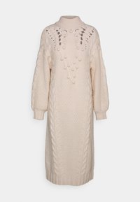 Abito maglione in beige con colletto alto. Presenta dettagli a trecce e pompon, maniche lunghe e una silhouette rilassata.