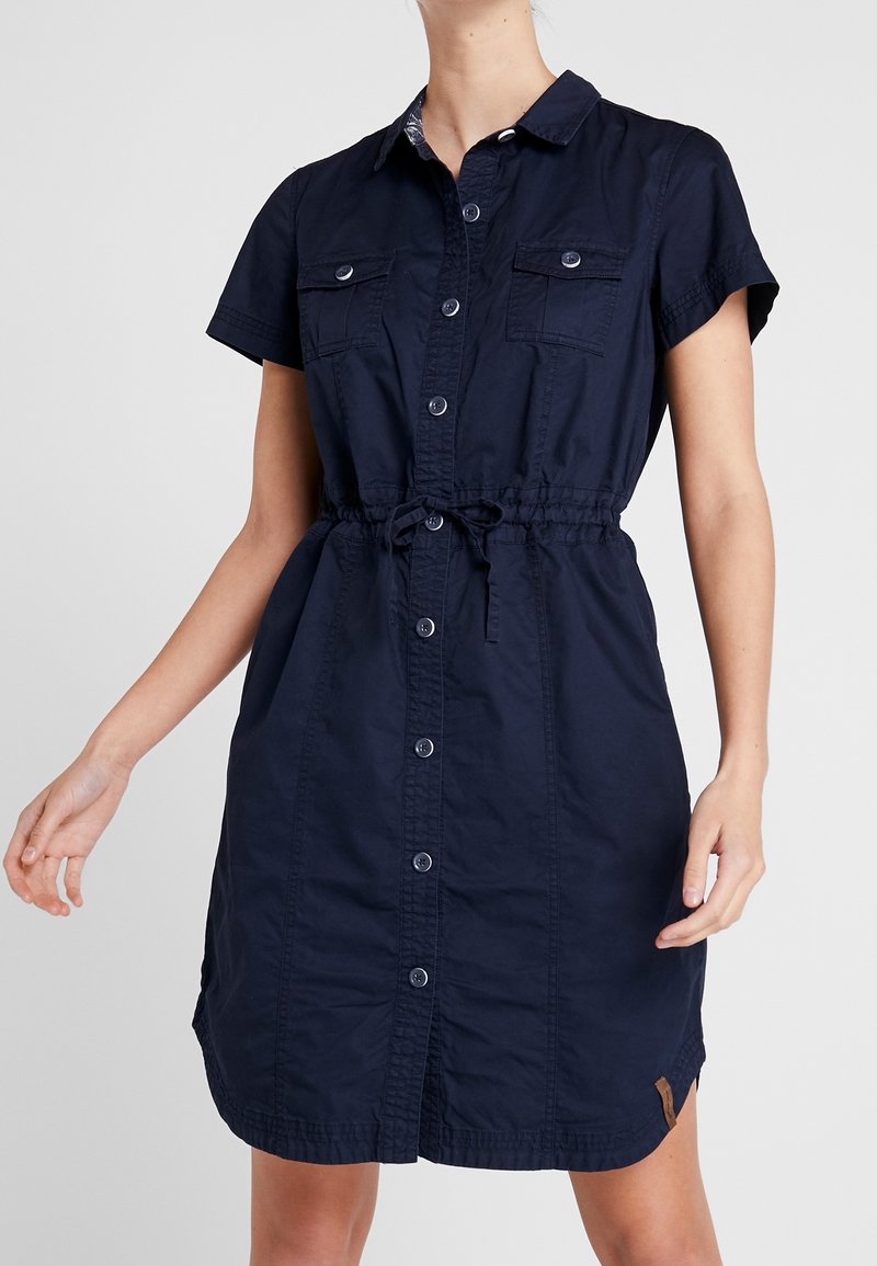 Femme portant une robe chemise bleu marine à manches courtes avec des boutons sur le devant et un cordon à la taille, debout devant un fond uni.