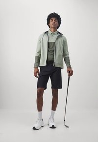 Světlězelená bunda na zip přes odpovídající polokošili, v kombinaci s černými šortkami a bílými golfovými botami. Drží golfovou hůl. Neutrální pozadí.