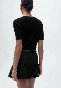 Schwarzes kurzärmliges Strickoberteil mit taillierter Silhouette, kombiniert mit einem schwarzen Röhrenrock mit Rüschen, getragen mit goldfarbenen Accessoires.