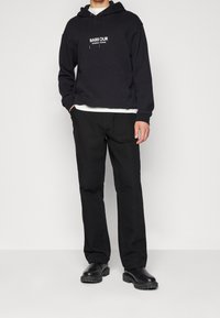 Černá mikina s kapucí s bílým nápisem "BARE OUR" na hrudi, doplněná černými kalhotami a černými botami, na jednobarevném pozadí.