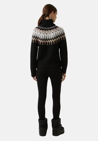 Vrouw die met haar rug naar de camera staat, gekleed in zwarte leggings, zwarte winterlaarzen en een zwarte coltrui met een wit en bruin patroon op de schouders.