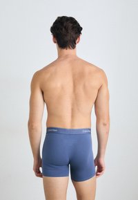 Kék pamut boxer alsó, szürke Calvin Klein derékkal, hátulról látható egy rövid hajú, póló nélküli személyen, látható részletek nélkül.