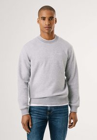 Sudadera gris de cuello redondo en tejido suave con puños y dobladillo de canalé; presenta un sutil bordado del logo en el pecho. Combinada con jeans azules.