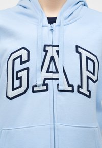 Sweat à capuche zippé bleu clair avec de grandes lettres brodées bleu marine et blanches "GAP" sur la poitrine, et cordons de serrage sur la capuche.