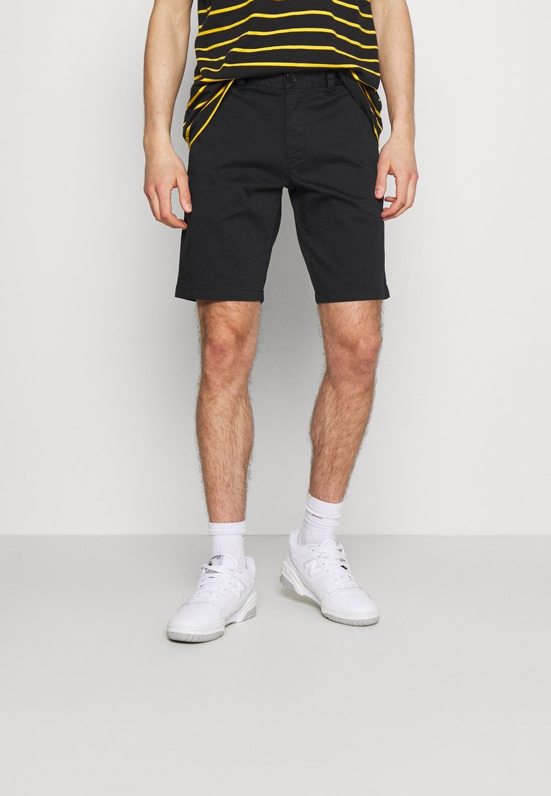 Homme portant un short noir, des baskets blanches et une chemise rayée noire et jaune, debout devant un fond uni.