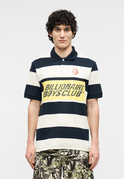 Νεαρός άνδρας με σγουρά μαλλιά, φορώντας μπλε σκούρο και λευκό ριγέ πόλο πουκάμισο με το κείμενο «Billionaire Boys Club» και cargo σορτς με σχέδια.