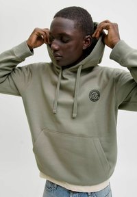 Oliivinvihreä huppari, joka on valmistettu pehmeästä kankaasta. Siinä on etutasku ja kiristysnyörillä varustettu huppu, sekä rintakehässä pyöreä logo.