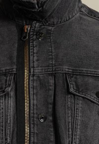 Närbild av en svart denimjacka med guldzipper, svarta tryckknappar, fickor med klaff och ståkrage.