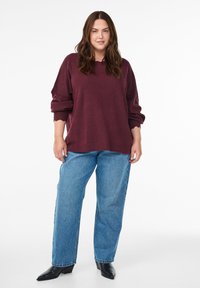 Burgunderfarbener, übergroßer Strickpullover mit gerippten Bündchen, kombiniert mit hellblauen, geraden Jeans und schwarzen, spitzen Ankle Boots.