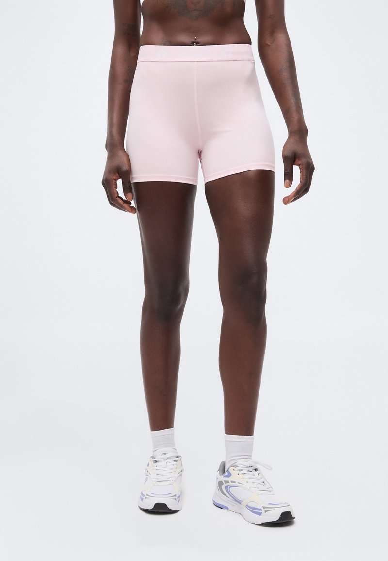 Άτομο που φοράει ανοιχτό ροζ στενά σορτς, λευκές κάλτσες και λευκά αθλητικά παπούτσια με μοβ και γκρι λεπτομέρειες, στέκεται μπροστά σε ένα απλό φόντο.