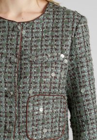 Grüne Tweedjacke mit strukturierten Oberflächen, runden Ausschnitt, Knöpfen und einer aufgenähten Tasche, die mit glänzenden Pailletten verziert ist.