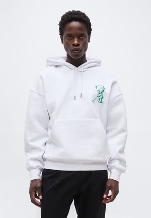 Νεαρός που φοράει λευκό φούτερ με κουκούλα με πράσινο γραφικό αρκουδάκι, κρατώντας ένα γκολφ κλαμπ, συνδυασμένο με μαύρο παντελόνι, στέκεται μπροστά σε ένα απλό φόντο.