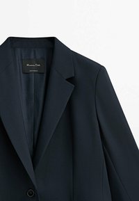 Blazer bleu marine foncé sur mesure avec col à revers cranté et un seul bouton visible, montrant la doublure intérieure et l'étiquette de la marque à l'intérieur du col.