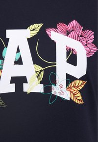 Námořnické tričko z bavlny s velkými bílými písmeny "AP" zdobené květinovými vzory v růžové, žluté a zelené barvě na levé straně.
