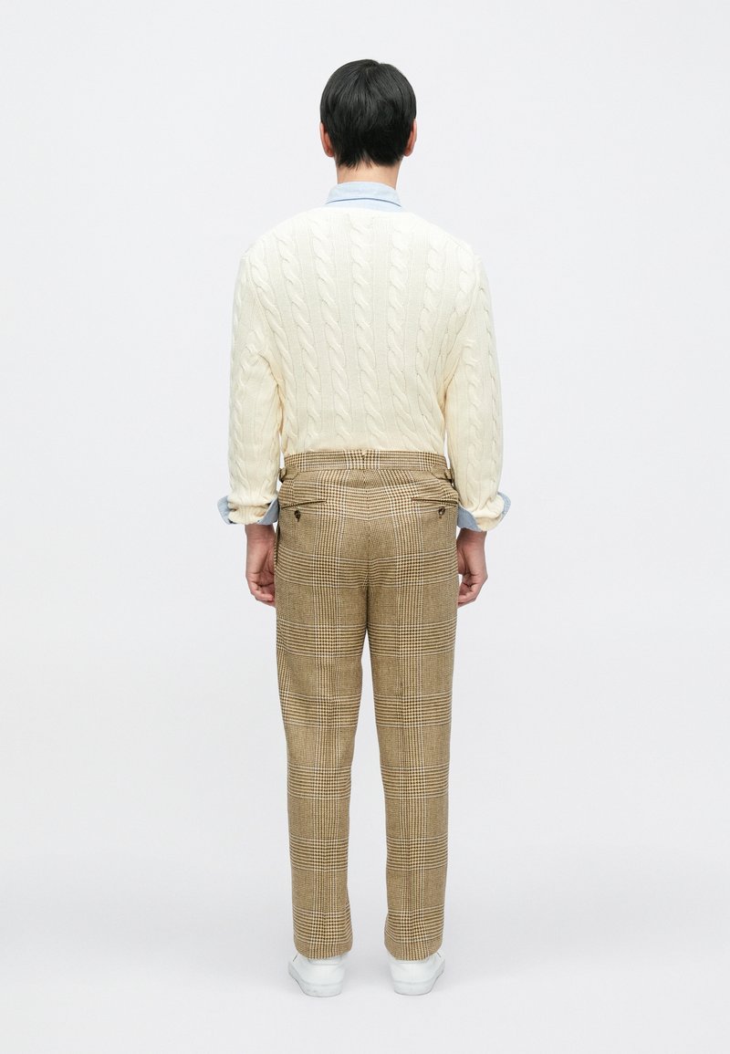 Cream cable-knit sweater, light blue shirt cuffs visible, paired with brown plaid trousers and white sneakers, viewed from the back.