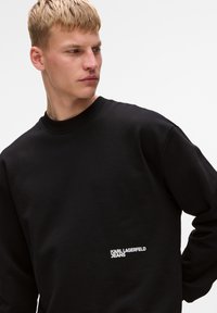 Felpa nera con colletto a girocollo, polsini a costine e logo ricamato "KARL LAGERFELD JEANS" sul lato sinistro. Tessuto morbido.