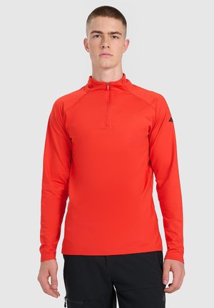 Młody mężczyzna noszący jaskrawoczerwony sportowy top z długim rękawem, z krótkim zamkiem z przodu oraz czarne spodnie, stojący na jednolitym tle.