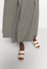 Une personne portant une longue jupe à motifs et des sandales à lanières tressées beiges se tenant sur un sol gris clair.