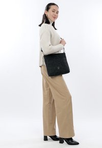 Femme portant un pantalon beige, un pull en tricot crème, des bottes à talons noires, tenant un sac à bandoulière noir sur fond blanc.