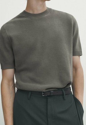 Personne portant un pull en maille vert olive à manches courtes rentré dans un pantalon vert foncé avec une ceinture noire, une main dans la poche.