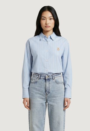 Giovane donna che indossa una camicia azzurra a righe verticali con piccoli bottoni e un piccolo emblema di orsetto, abbinata a jeans a lavaggio chiaro.