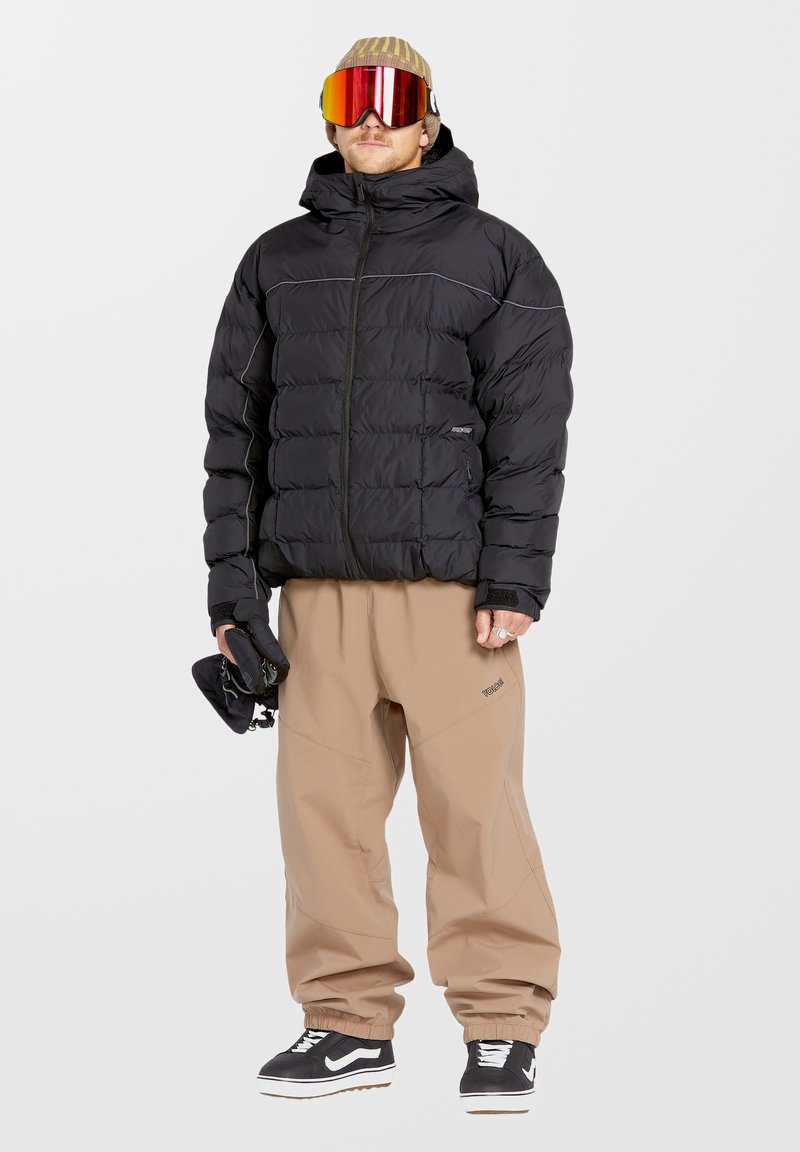 Zwarte gewatteerde jas met een capuchon, voorzien van een ritssluiting aan de voorkant en grijze accenten. Beige broek gemaakt van een zachte, flexibele stof. Witte sneakers.