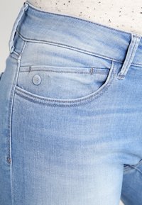 Světle modré džíny z denimu s elegantním designem, mírným vyblednutím, přední kapsou a hladkou texturou. Viditelné detaily knoflíku a prošívání.