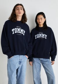 Sudaderas de color azul marino con el logo "TOMMY JEANS" en blanco, cuello redondo, corte holgado, combinadas con jeans de color azul claro, textura de tela casual.