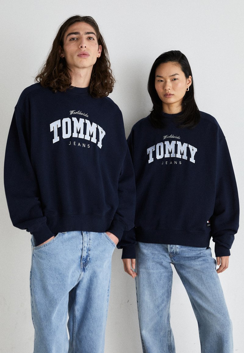 Sudaderas de color azul marino con el logo "TOMMY JEANS" en blanco, cuello redondo, corte holgado, combinadas con jeans de color azul claro, textura de tela casual.