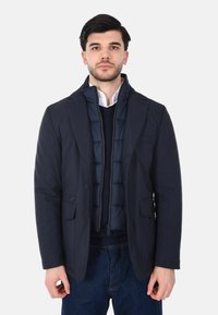 Giacca ibrida blu navy con imbottitura interna, chiusura frontale con bottoni e due tasche frontali. Tessuto liscio con colletto e polsini strutturati.