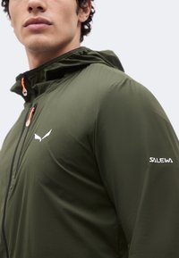 Νέος άνδρας φοράει μπουφάν Salewa ελιάς με κουκούλα, ορατά λογότυπα στο στήθος και στο μανίκι, κοιτάζοντας ελαφρώς πλάγια.