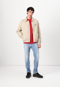 Beige jakke med elastiske mansjetter over rød hettegenser, kombinert med lyseblå jeans og svarte sneakers. Merke på venstre side av jakken.