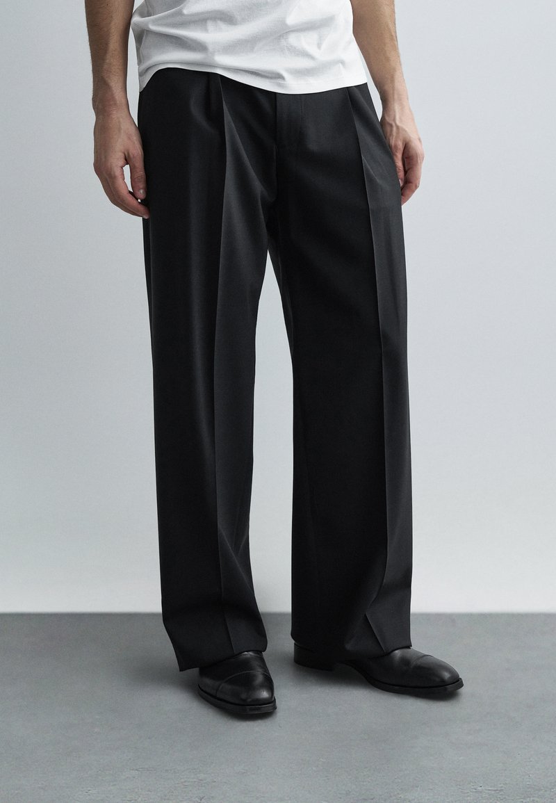 Juodos, klostuotos kelnės, laisvo silueto, pagamintos iš lygaus audinio. Dėvimos su juodais batais, derinamos su paprasta balta marškinėlių palaidine.