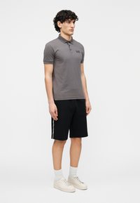 Grå bomulds-polo med korte ærmer, sorte shorts med sidelommer, og beige sneakers. Modell står mod en ensfarvet baggrund.