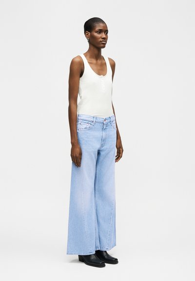 Naine, kes seisab ja kannab valget varrukateta toppi, helerohelisi laia säärega teksapükse ja musti pahkluu saapaid tavalise heledat tausta ees.