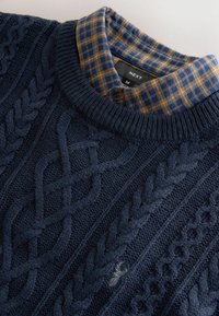 Marineblå strikket sweater med kabelmønstre, der har en krave i brune og blå tern. Materialet virker blødt med en tekstureret overflade.