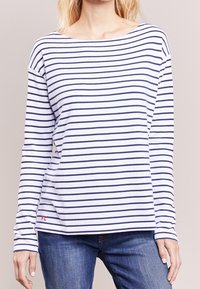 Frau trägt ein weißes Langarmshirt mit horizontalen marineblauen Streifen und blaue Jeans, steht gegen einen schlichten beigen Hintergrund.