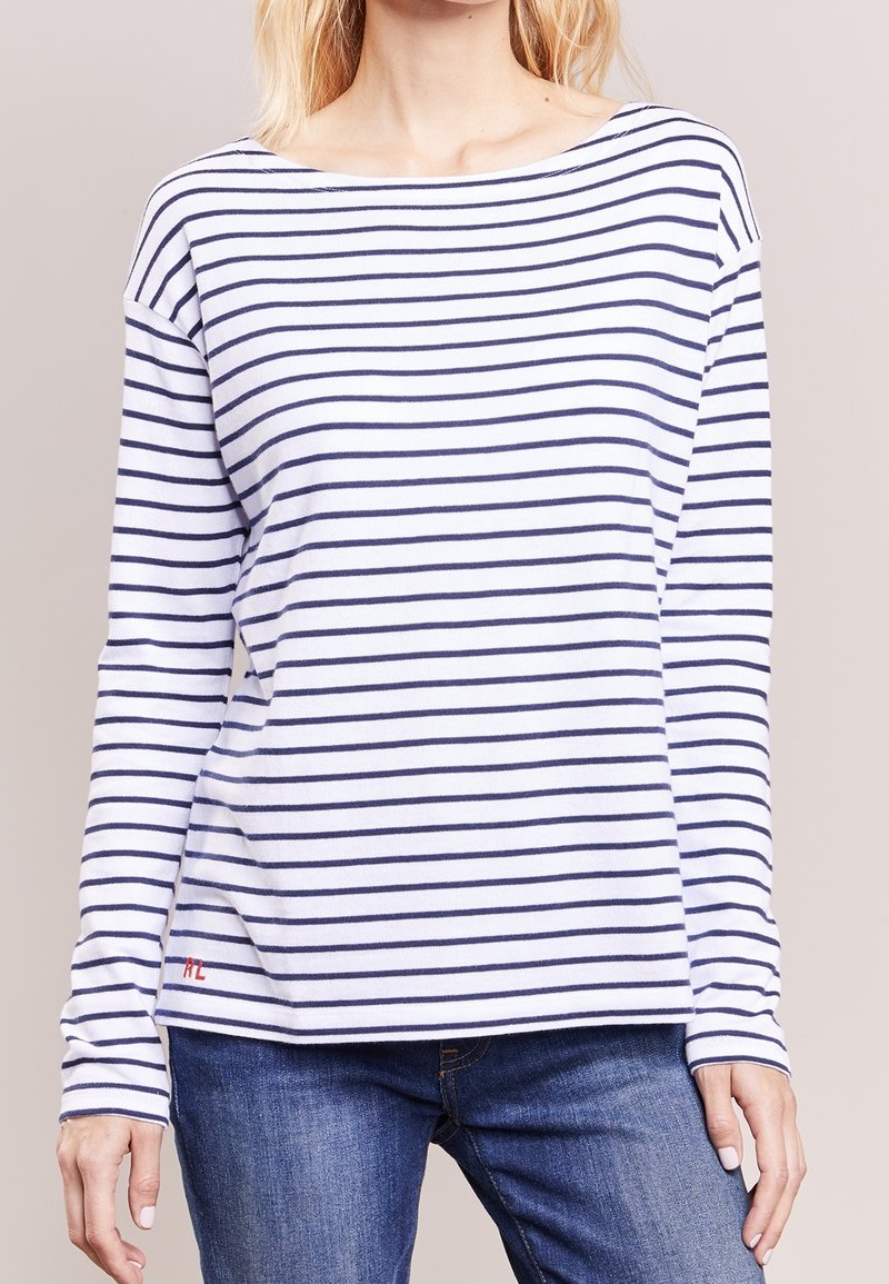 Frau trägt ein weißes Langarmshirt mit horizontalen marineblauen Streifen und blaue Jeans, steht gegen einen schlichten beigen Hintergrund.
