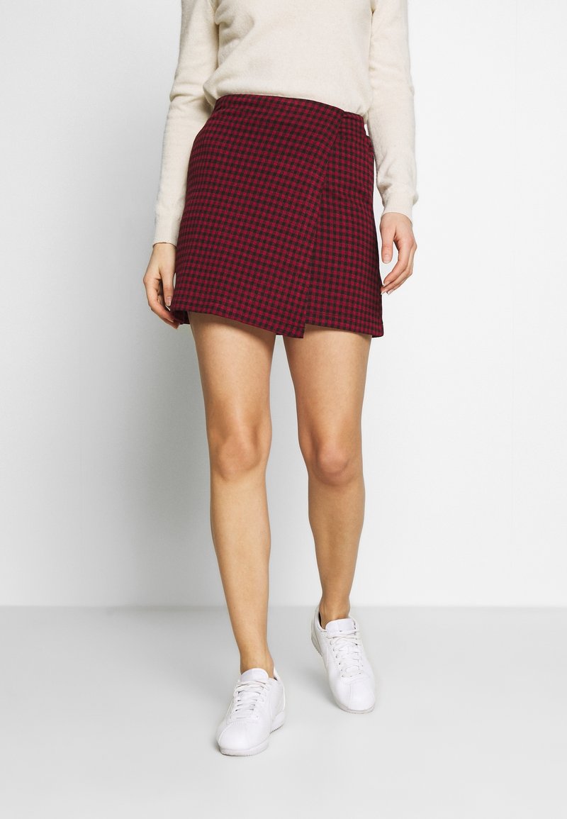 Femme portant une jupe portefeuille à carreaux rouges et noirs, un pull blanc et des baskets blanches, debout devant un fond clair uni.
