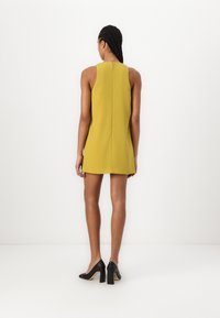 A sleeveless yellow shift dress with a high neck and back zipper, paired with black heels, featuring a straight cut and minimal detailing.