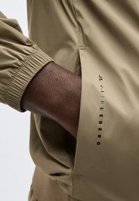 Σκούρο χέρι μέσα στην τσέπη ενός μπεζ σακακιού με ελαστικό μανσέτι, με μαύρο λογότυπο "J.LINDEBERG" στην άκρη της τσέπης.