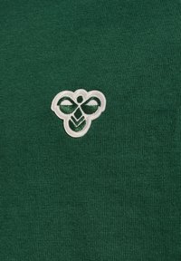 Camisa de algodón verde con un logo de abeja bordado en blanco en el lado izquierdo. La tela tiene una textura suave con un diseño ajustado.