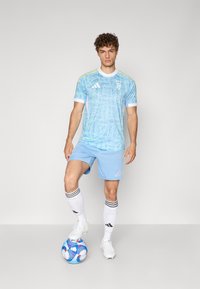 Lyseblå Adidas-jumper med hvite og gule detaljer, med abstrakte mønstre. Kombinert med matchende blå shorts og hvite sokker.