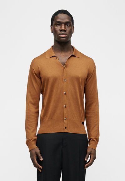 Άνδρας που φοράει καφέ μακρυμάνικη ζακέτα με κουμπιά και γιακά, καθώς και μαύρο παντελόνι, στέκεται μπροστά σε απλό ανοιχτόχρωμο φόντο.