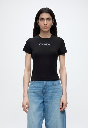 Νεαρή γυναίκα με μακριά σκούρα μαλλιά, φορώντας μαύρο t-shirt Calvin Klein και ανοιχτό μπλε ψηλόμεσα τζιν, στέκεται μπροστά σε απλό φόντο.