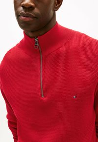 Pull rouge en tricot avec un col montant et une fermeture à demi-zip. Présente un motif texturé et un petit logo brodé sur la poitrine.
