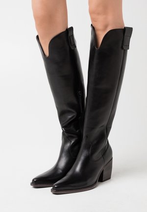 Bottes en cuir noires noires montantes jusqu'aux genoux avec bouts pointus et talons carrés portées sur des jambes nues sur fond blanc.