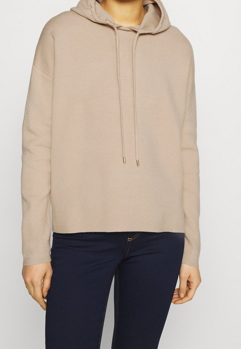 Beige Kapuzenpullover mit lockerer Passform, ausgestattet mit einem Kordelzug mit goldenen Akzenten und gerippten Bündchen. Kombiniert mit dunkelblauen Jeans.