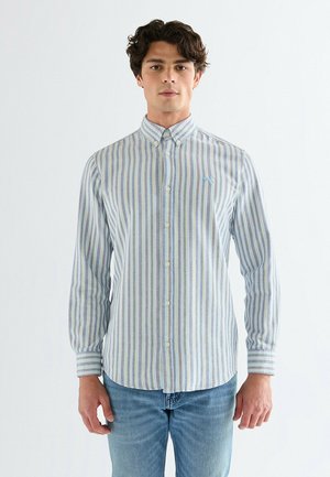 Mężczyzna w zapinanej na guziki, długorękawowej koszuli w pionowe, wielokolorowe paski i niebieskich dżinsach, stojący na jednolitym tle.